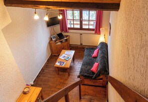 Un salon chaleureux avec un canapé en bois, une table basse et un téléviseur. Il y a une fenêtre avec des rideaux, un radiateur et une lampe.