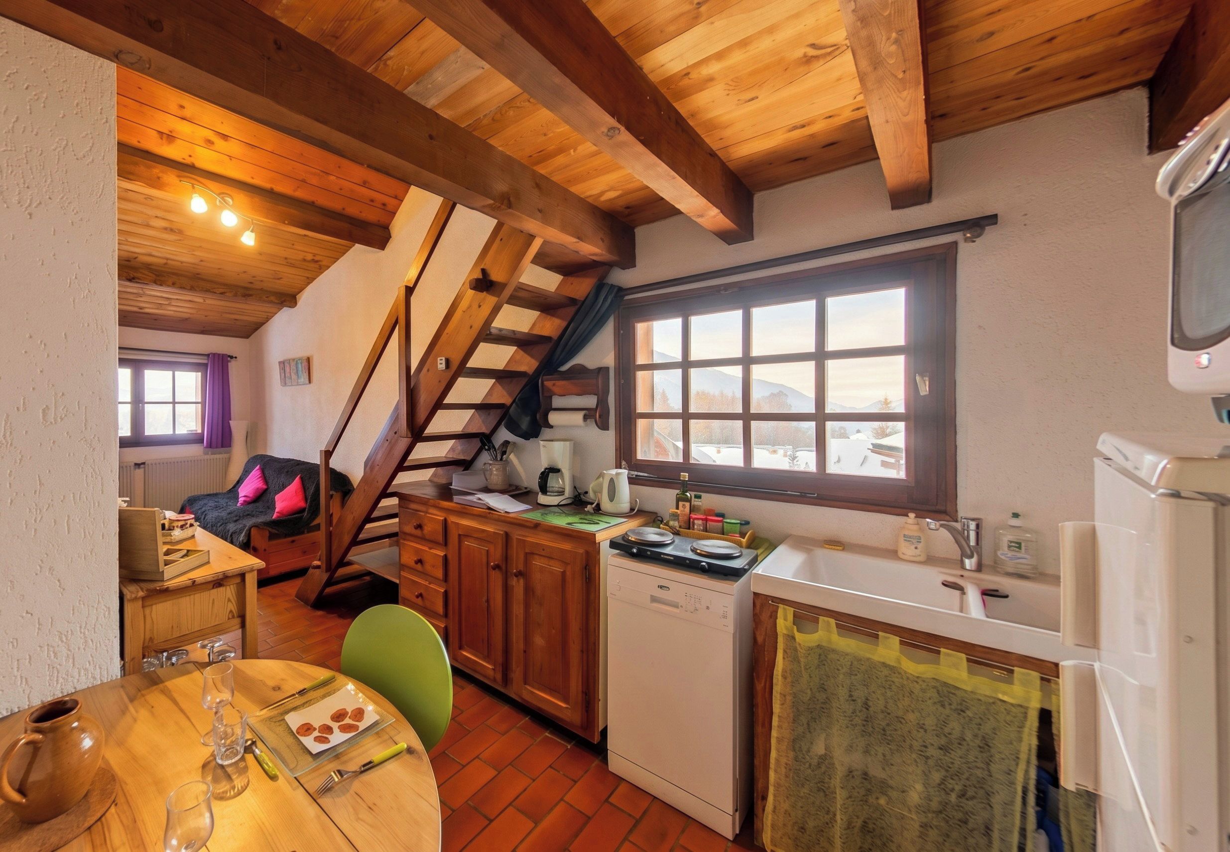 Une cuisine chaleureuse avec des poutres en bois, un coin repas avec une table dressée pour deux personnes, et une fenêtre donnant sur un paysage enneigé.
