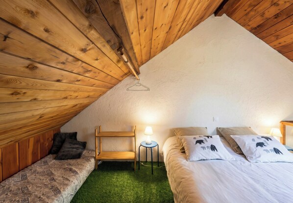 Une chambre mansardée chaleureuse, avec un plafond en pente, un lit superposé, une petite table avec une lampe et un sol recouvert d’un tapis d’herbe artificielle verte.