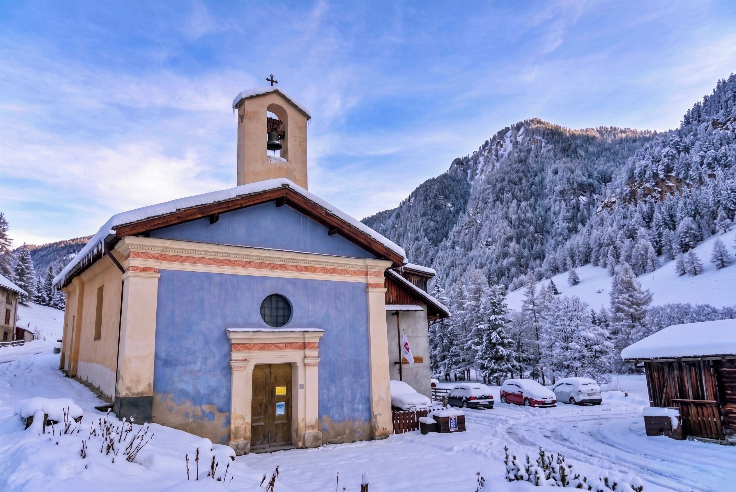 A small church with a bell tower, surrounded by snow-covered trees and mountains.