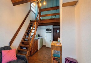 Un ambiente compatto con una scala in legno che conduce a un livello superiore, una zona cottura con elettrodomestici essenziali e un piccolo angolo salotto con un divano e una poltrona.
