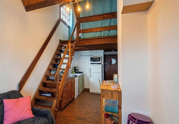 Ein kompakter Wohnbereich mit einer Holztreppe zu einer erhöhten Ebene, einer Küchenzeile mit einfachen Geräten und einer kleinen Sitzecke mit Sofa und Sessel.