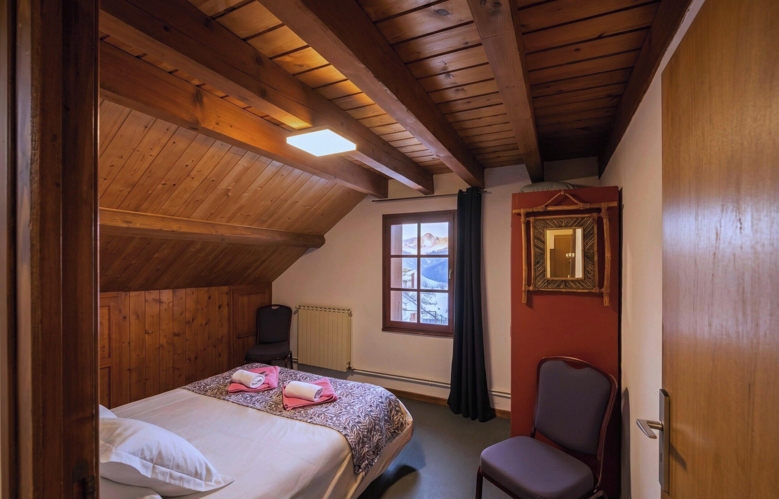A bedroom with a wooden ceiling, a bed with pillows, a chair, a window with a view, and a radiator.