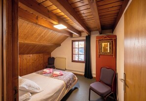 A bedroom with a wooden ceiling, a bed with a patterned bedspread, a chair, a window with a view of mountains, and a radiator.