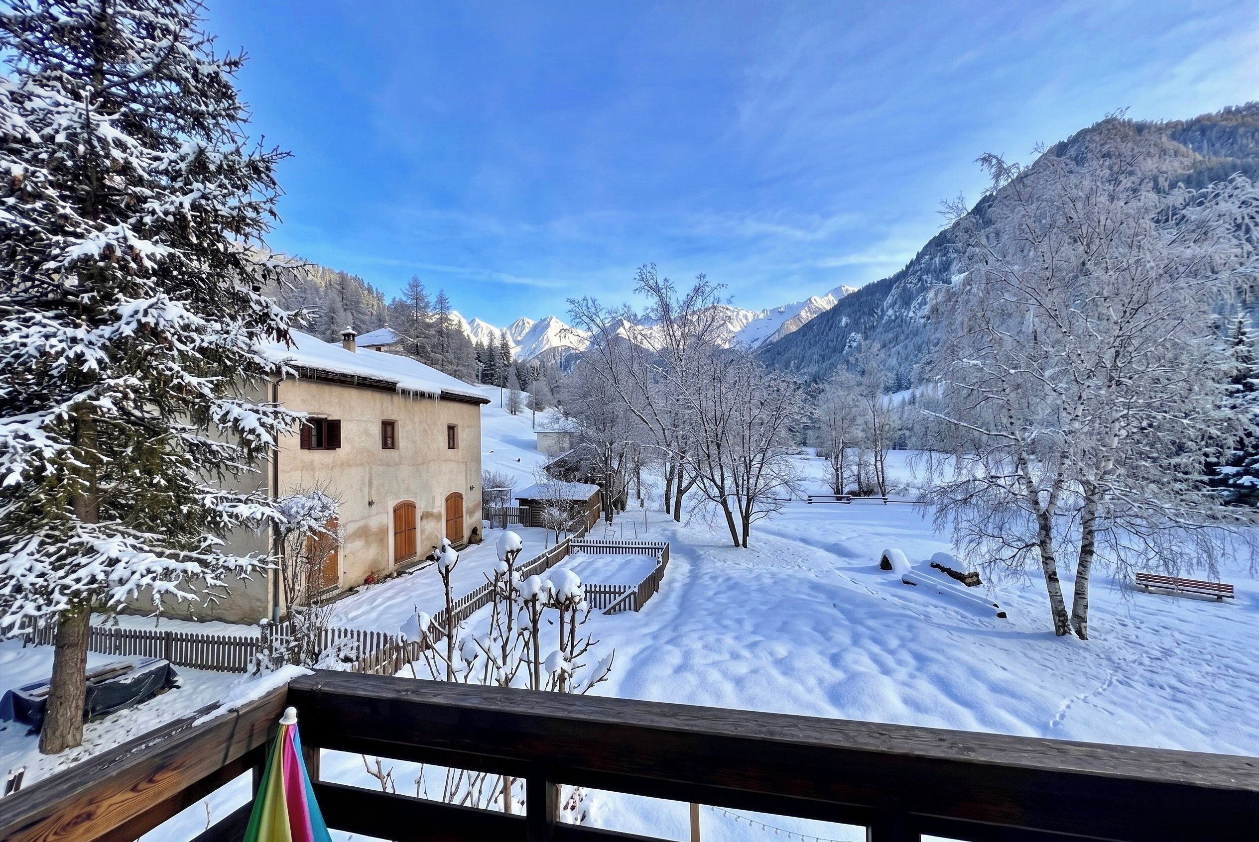 Un village enneigé avec un bâtiment en bois, un sentier recouvert de neige et des montagnes en arrière-plan.
