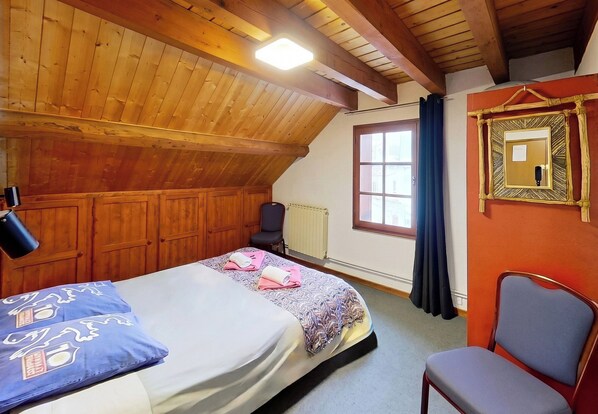 Une pièce avec un plafond en bois, un lit avec une literie à motifs, une chaise, un radiateur et une fenêtre avec des rideaux.