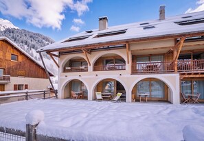 A multi-story chalet with a snow-covered roof, wooden balconies, and a ski slope in the background.
