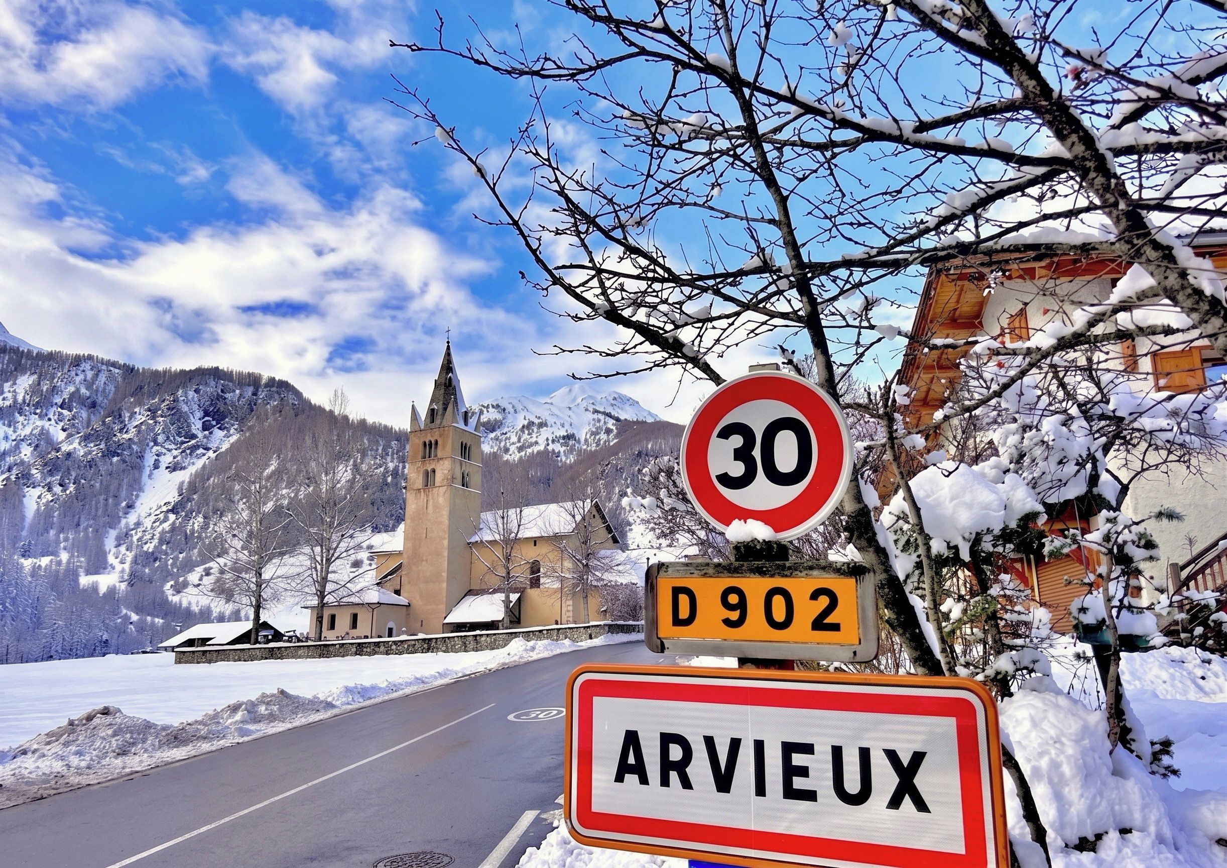 A road sign with '30' and 'D902' indicating a speed limit and a route number, with 'ARVIEUX' below it, set against a backdrop of a snow-covered landscape with a church and mountains.