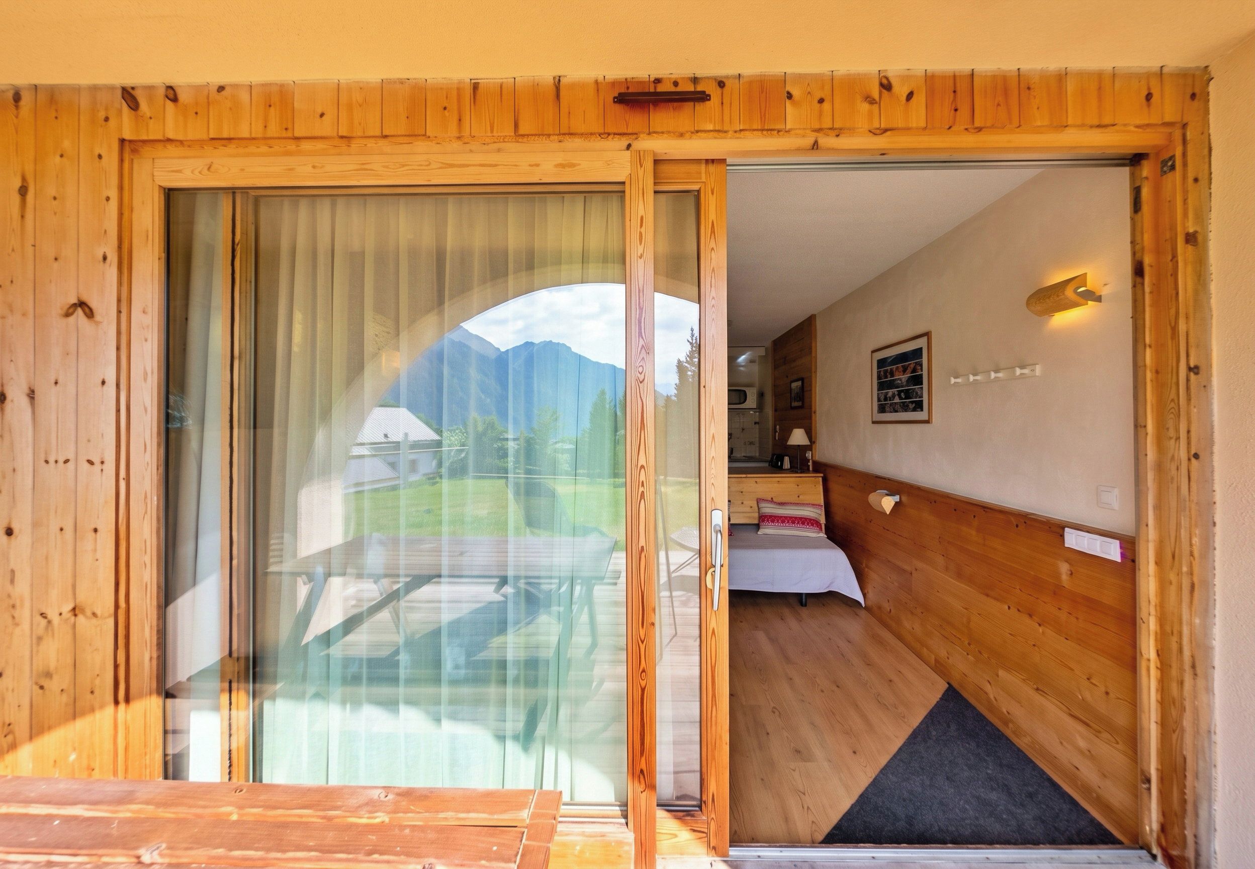 A wooden interior with a glass door leading to a balcony, a bed with a pillow, a wall-mounted light, and a framed picture.