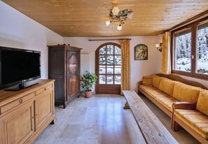 Ein gemütliches Wohnzimmer mit Fernseher, Holzmöbeln, einer Topfpflanze, einer Bank und einem großen Fenster mit Blick auf schneebedeckte Bäume.