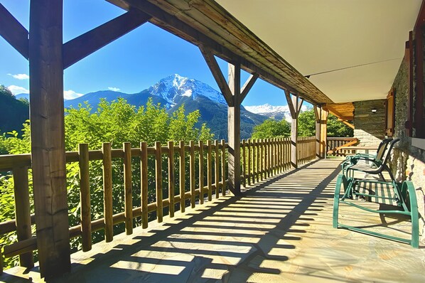 Une terrasse en bois offrant une vue sur des montagnes enneigées, un mur en pierre et un banc en métal vert.