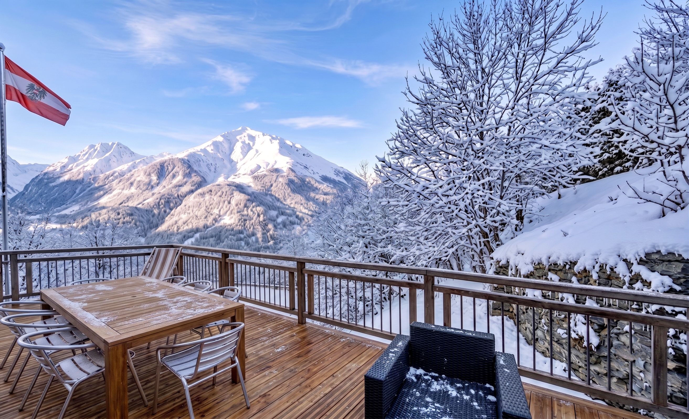 A deck with a table and chairs, overlooking snow-covered mountains and a flag.