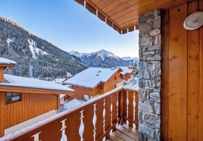 A wooden balcony with a stone wall, overlooking a snowy mountain landscape and a chalet.