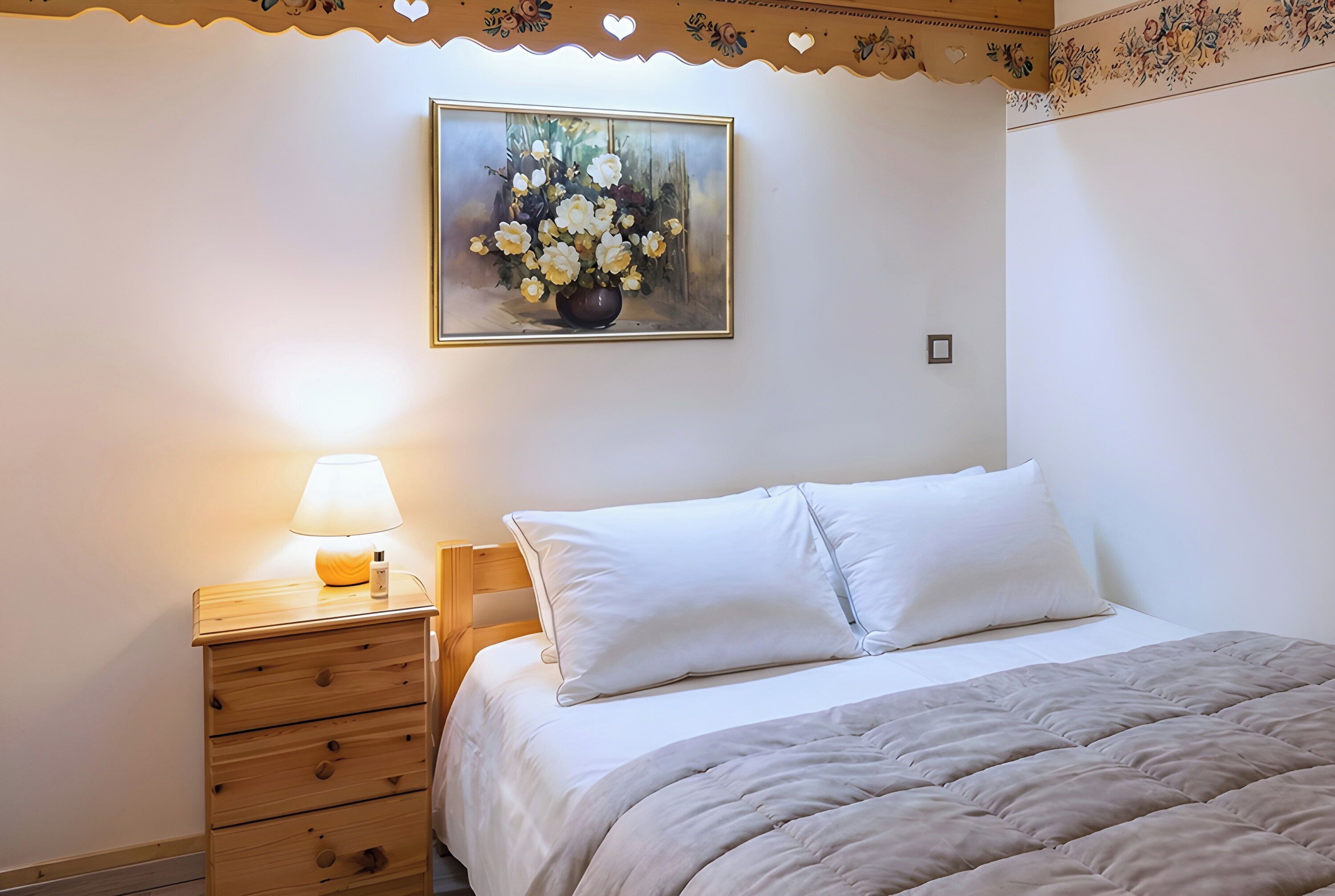 Een slaapkamer met een bed, een nachtkastje met een lamp, een houten dressoir en een lijst met een bloemistisch schilderij aan de muur.