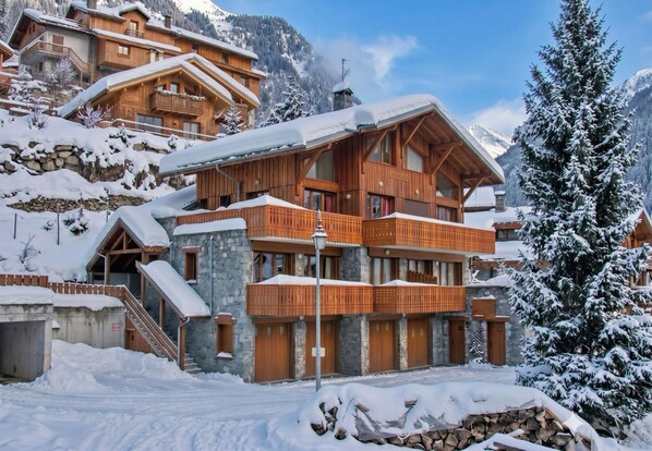 A multi-story wooden chalet with balconies, set against a snowy mountain backdrop.