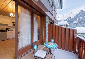 Un balcon avec vue sur des montagnes enneigées, une table à manger avec des chaises et un coin cuisine équipé d’une cuisinière et de placards.