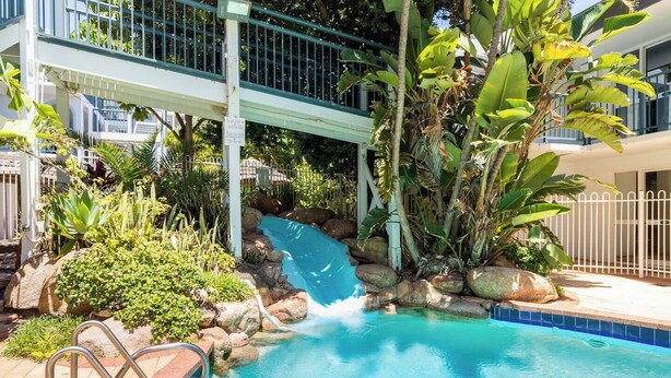 A pool with a waterfall feature, surrounded by tropical plants and rocks. - Coastal Beach Retreat Scarborough Pool & Slide! (Scarborough)