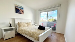 A bedroom with a bed, a nightstand, a lamp, a painting on the wall, and a view of the outside through a window. - Coastal Beach Retreat Scarborough Pool & Slide! (Scarborough)