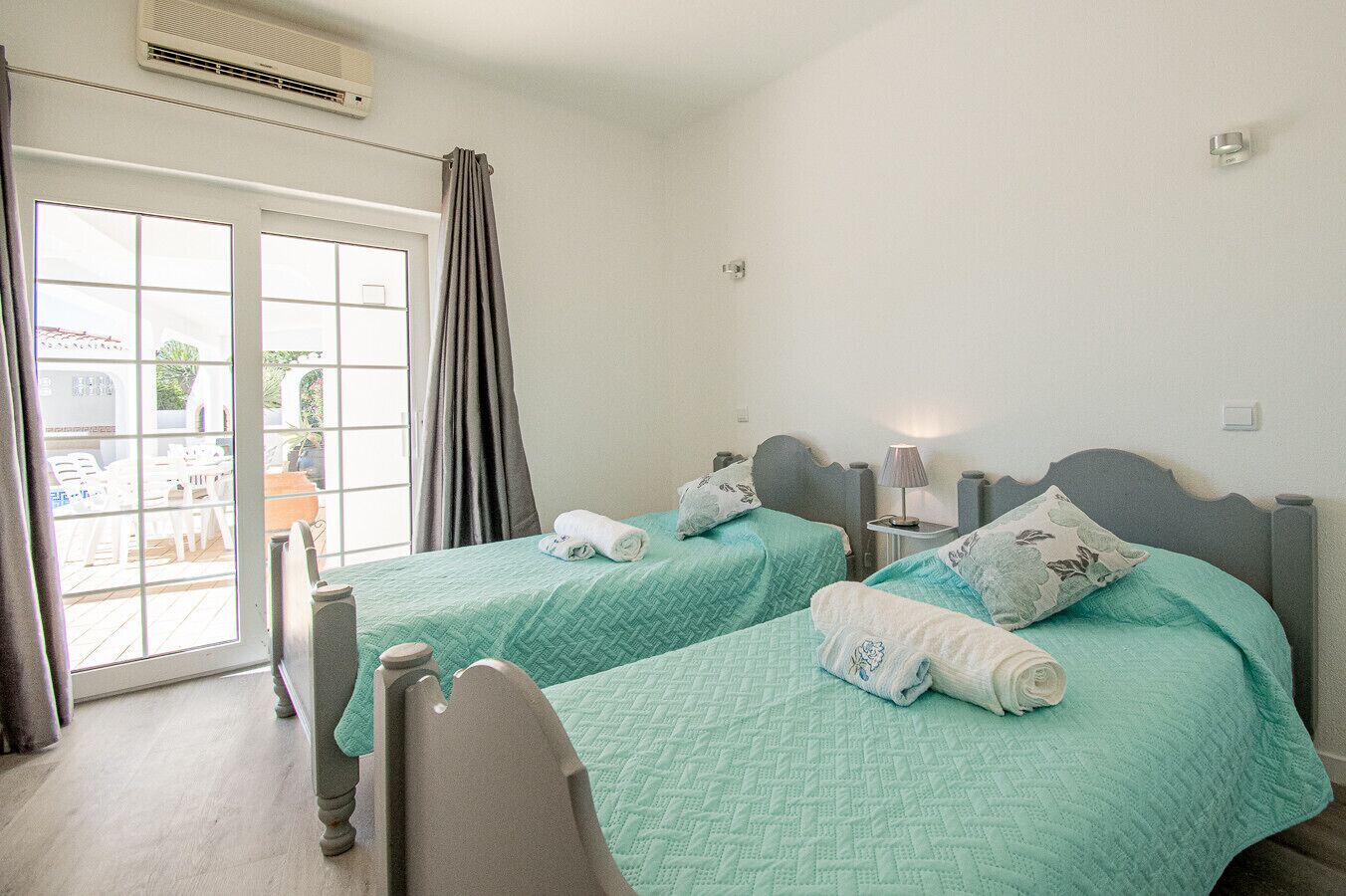 A room with two beds, a window with curtains, a small table with a lamp, and a view of an outdoor area with chairs.