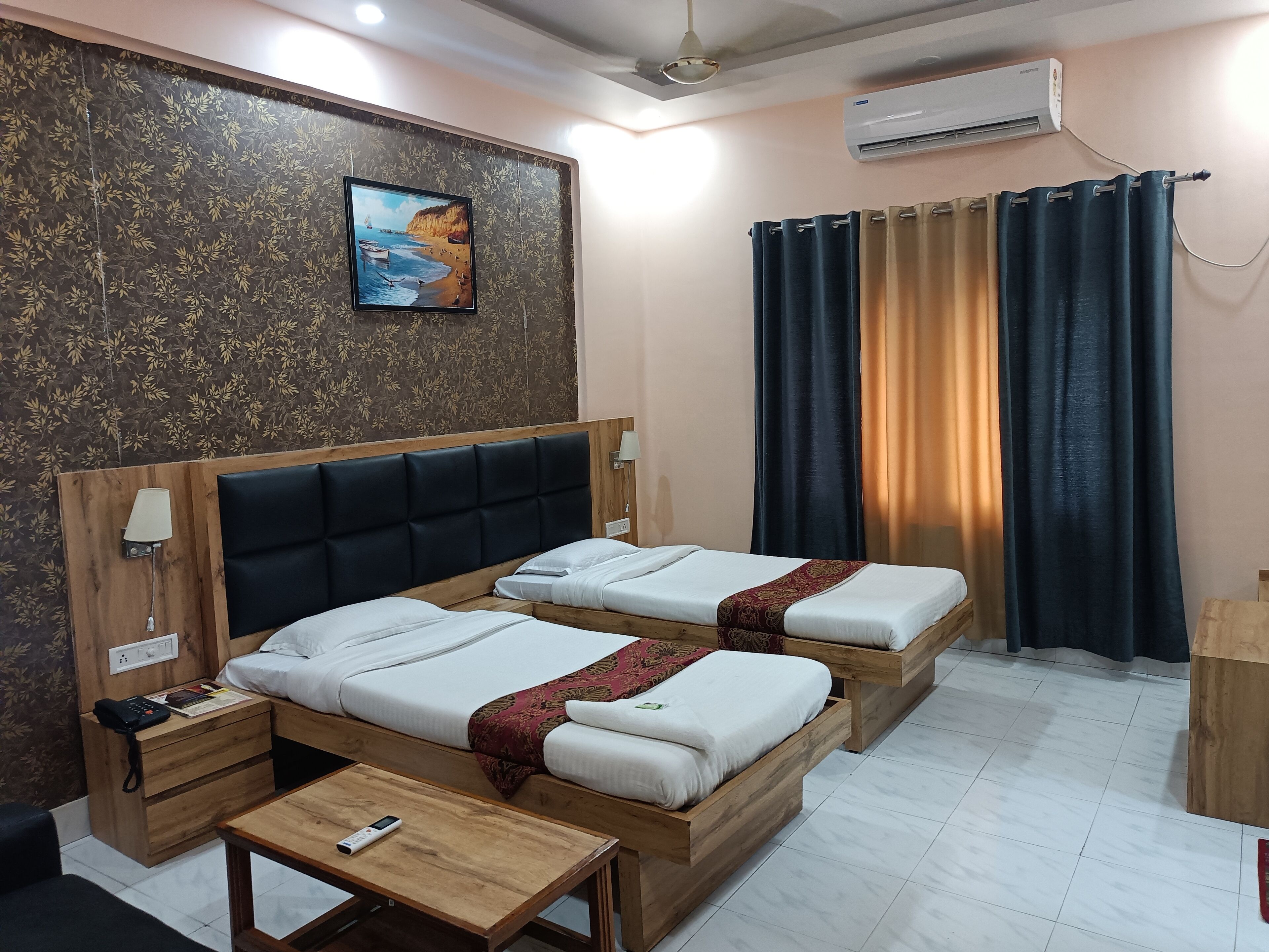 A hotel room with two beds, a wooden headboard, a bedside table with a lamp, a wall-mounted air conditioner, and a painting of a beach scene.