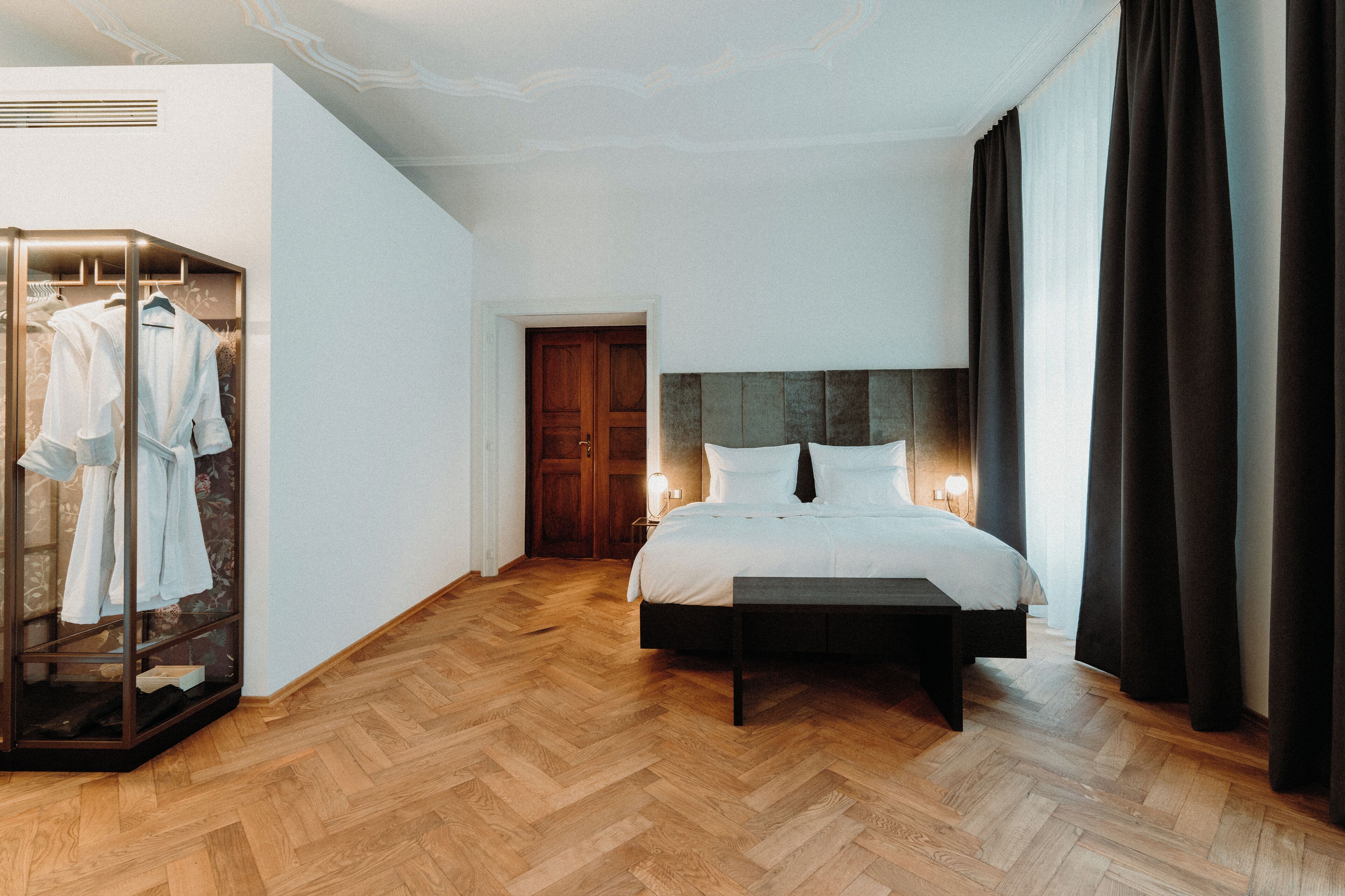 Una camera da letto con pavimento in parquet a spina di pesce, un letto con testiera scura, un comodino e una cabina armadio con abiti appesi.
