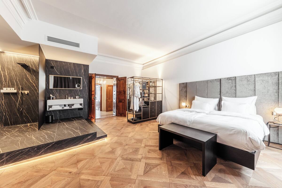 A modern hotel room with a large bed, a built-in bench, a vanity with a mirror, and a walk-in closet.