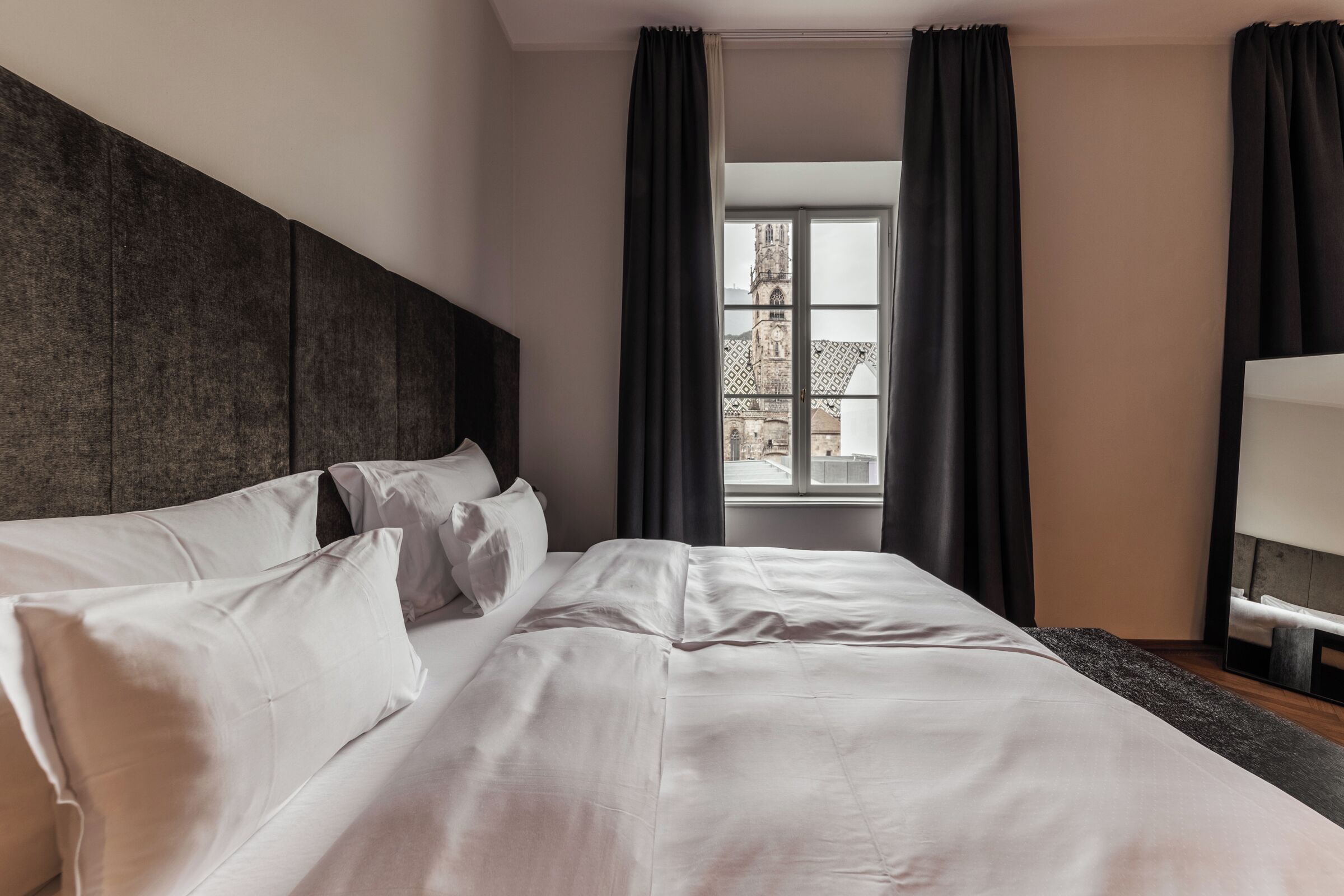 A neatly made bed with white linens and pillows, a dark headboard, and a window with a view of a building.