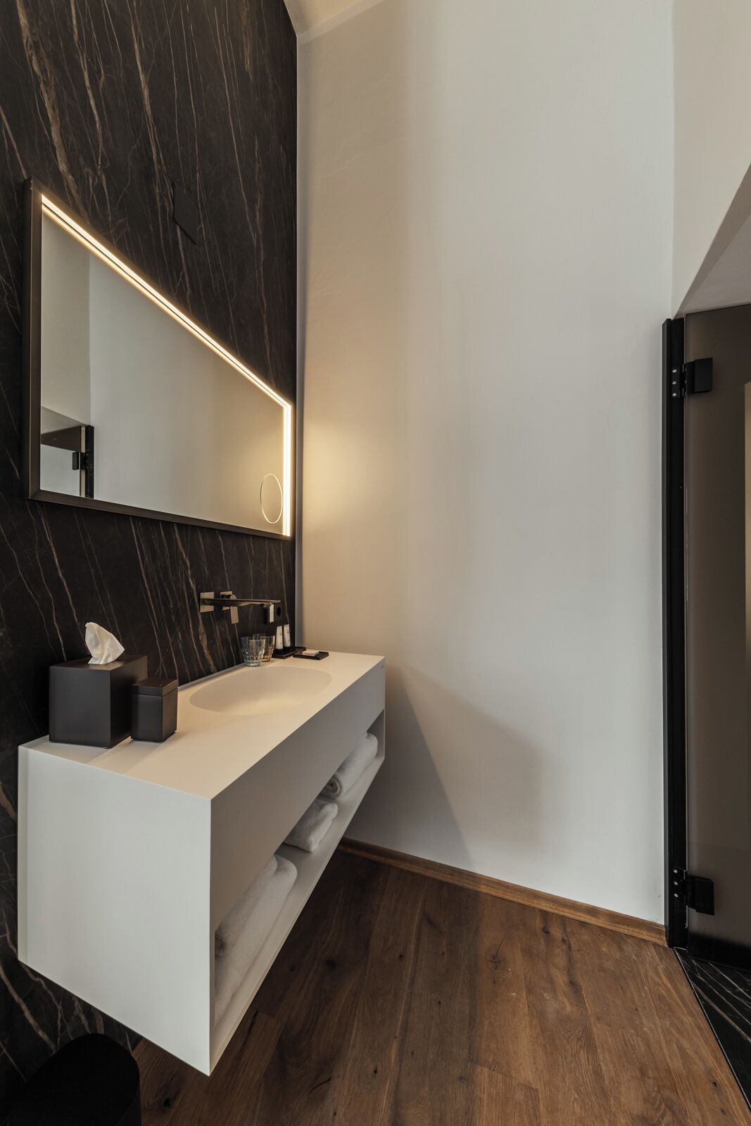 A modern bathroom with a marble wall, a large mirror, a white vanity, and a wooden floor.