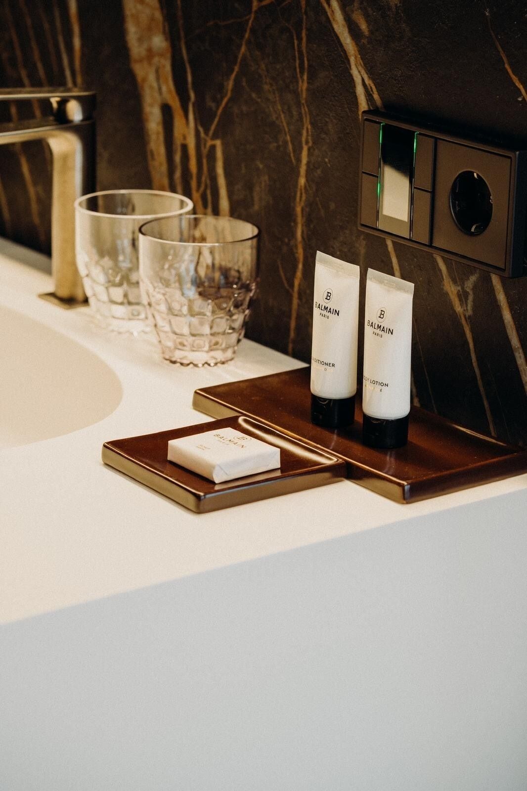 A bathroom vanity with a marble backsplash, a gold faucet, and two Balmain lotion tubes on a tray.