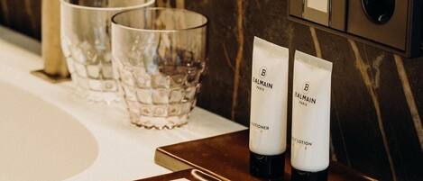 A bathroom vanity with a marble backsplash, a gold faucet, and two Balmain lotion tubes on a tray.
