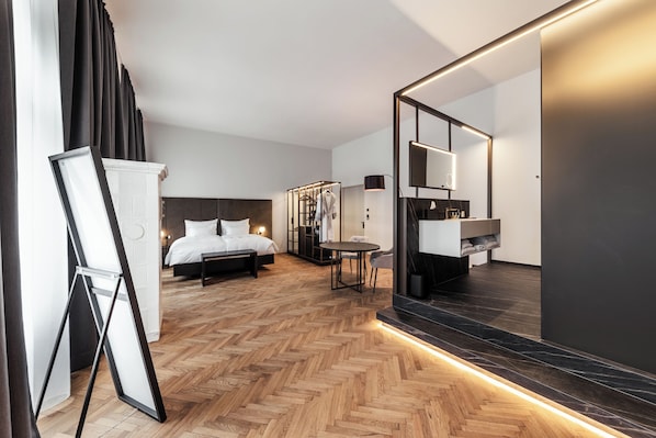 Una habitación de hotel moderna con piso de madera en patrón de espiga, una cama con sábanas blancas, una mesita de noche, una pequeña mesa redonda con una silla, un espejo grande y un armario de tono oscuro.