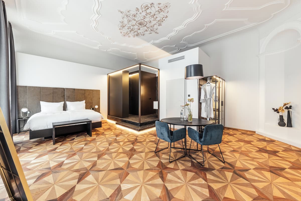 A modern hotel room with a geometric patterned floor, a bed with white linens, a bedside table, a mirror, a round table with two chairs, and a ceiling with decorative elements.