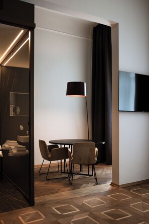 A modern hotel room with a round table, two chairs, a floor lamp, a flat-screen TV, and a large mirror.