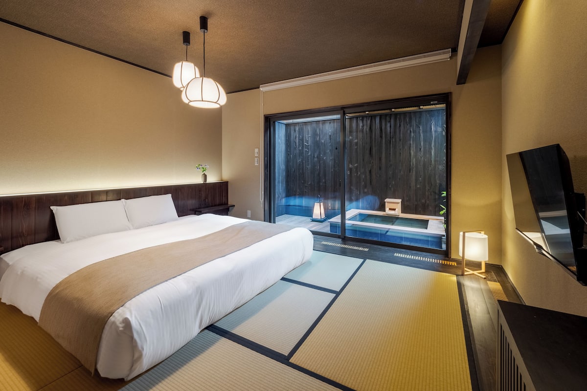 Ein Hotelzimmer mit einem großen Bett, einer Glastür zu einem Balkon, einem kleinen Tisch mit Lampe und einem Fernseher auf einem Schrank.