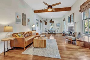 A spacious living room with a wooden floor, a ceiling fan, a fireplace, and a dining area with chairs and a table.