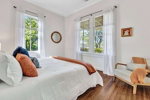 A bedroom with a bed, two windows, a chair, and a wall-mounted mirror.
