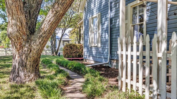A house with a wooden picket fence, a pathway leading to the front door, and a large tree in the foreground.