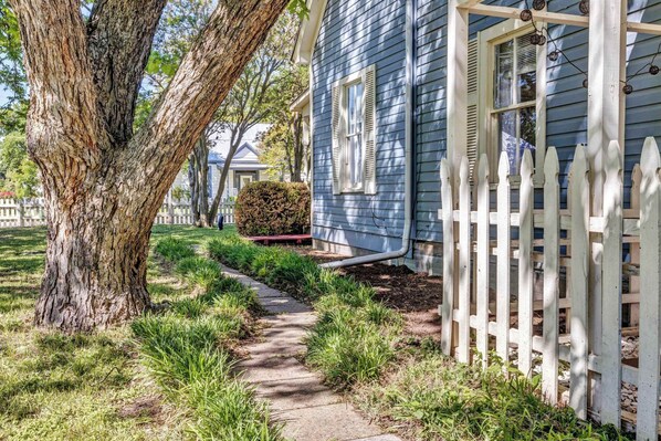 A pathway leading to a house with a white picket fence and a tree on the left.