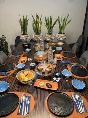 Una mesa de comedor dispuesta con platos, cubiertos y diferentes platos, rodeada de sillas y plantas en macetas.