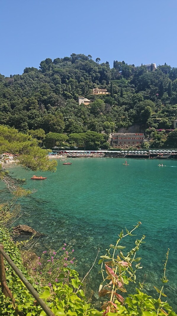 Uma área costeira com águas turquesa cristalinas, uma praia com guarda-sóis e edifícios numa encosta.
