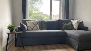 Uma sala de estar moderna com um sofá modular cinza, uma mesinha de apoio com uma planta e uma grande janela com cortinas.