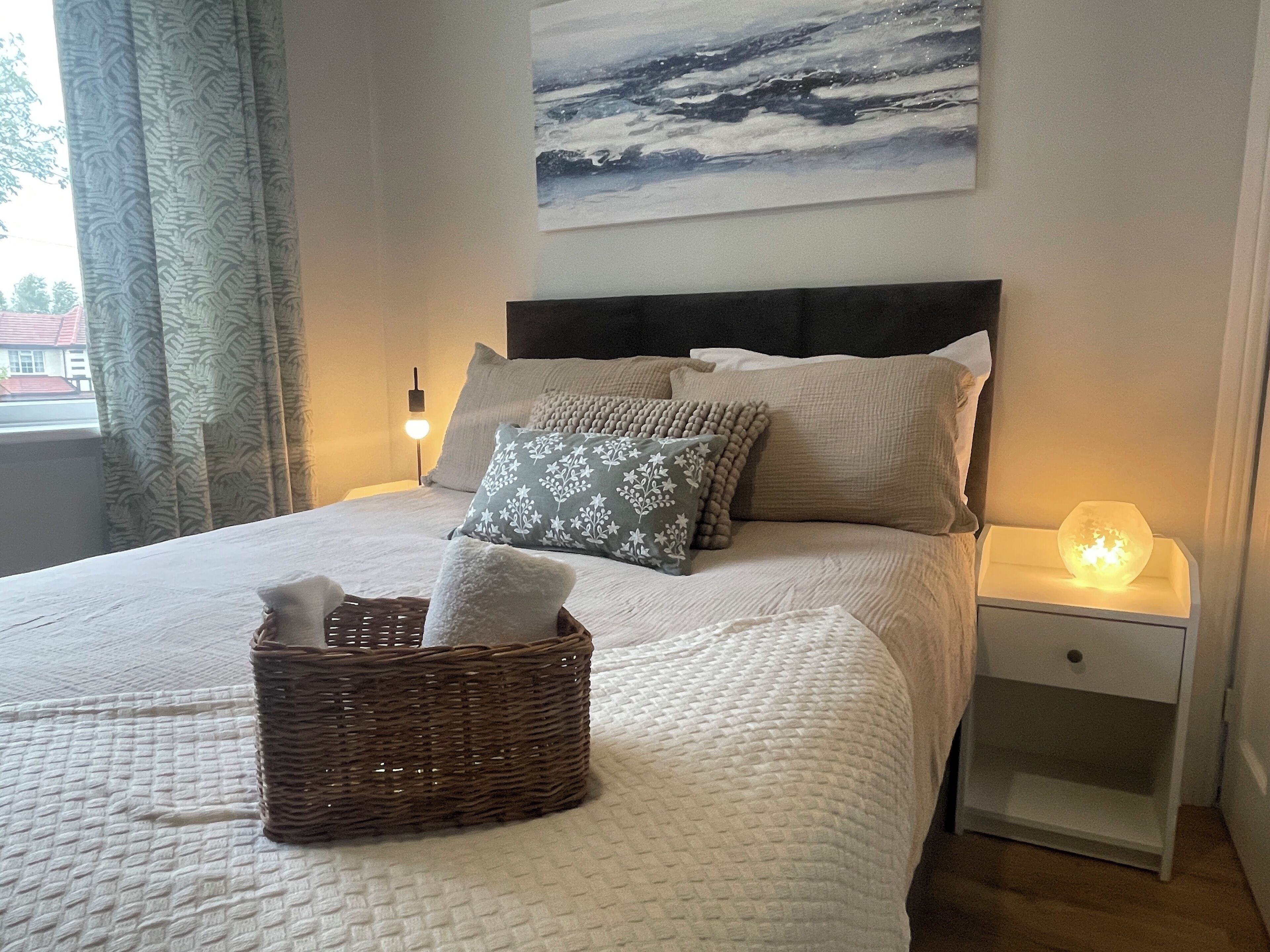 A neatly made bed with a basket of towels, a nightstand with a lamp, and a painting on the wall.