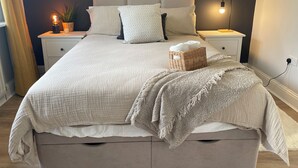 Cama bem arrumada com cabeceira branca, mesa de cabeceira com abajur e pequeno cesto na cama.