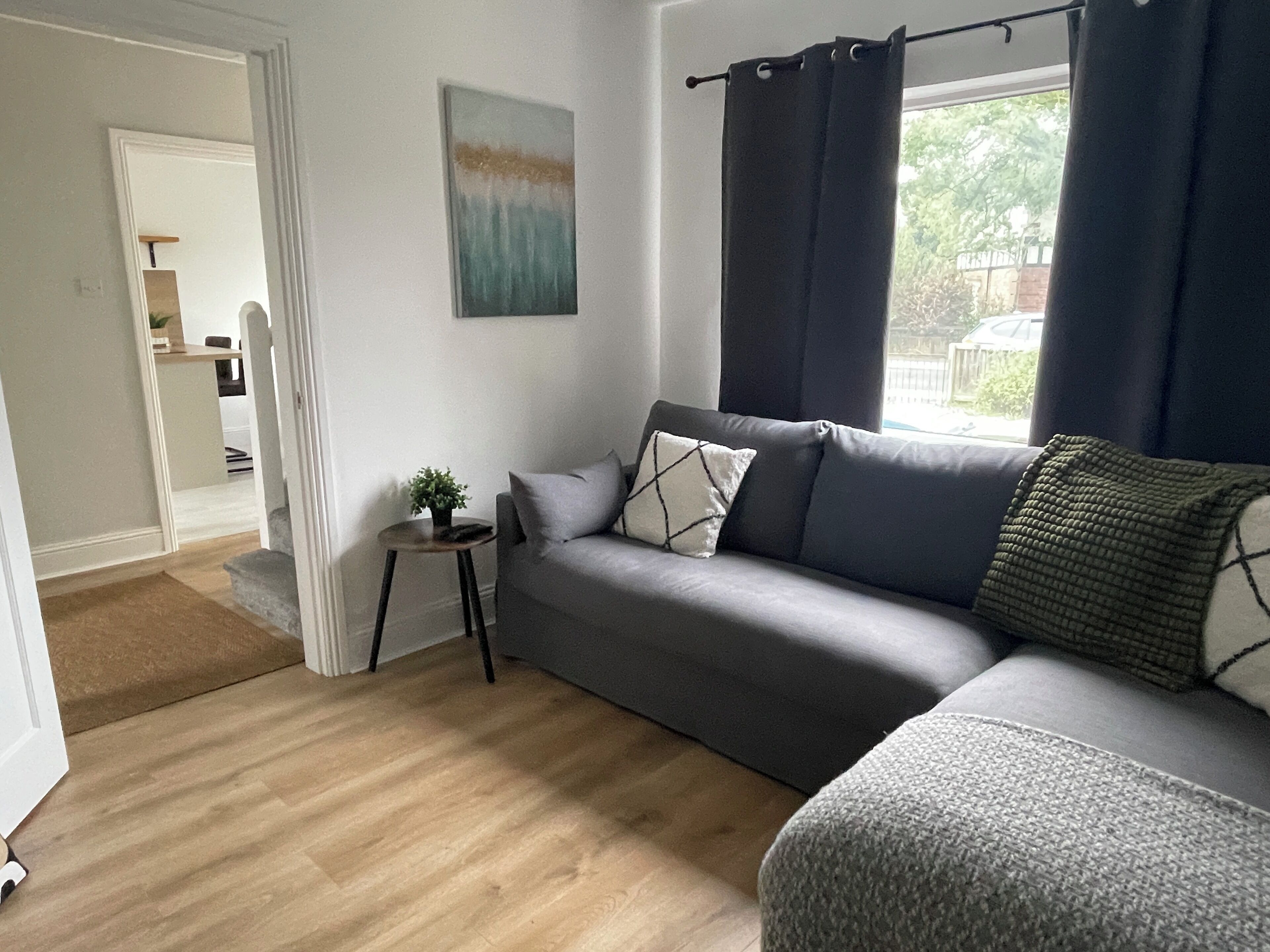 A modern living room with a grey sofa, a small side table with a plant, and a window with dark curtains.
