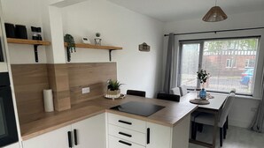 Cozinha moderna com bancadas de madeira, armários brancos e fogão embutido. Há uma área de jantar com mesa e cadeiras, e uma janela com persianas.