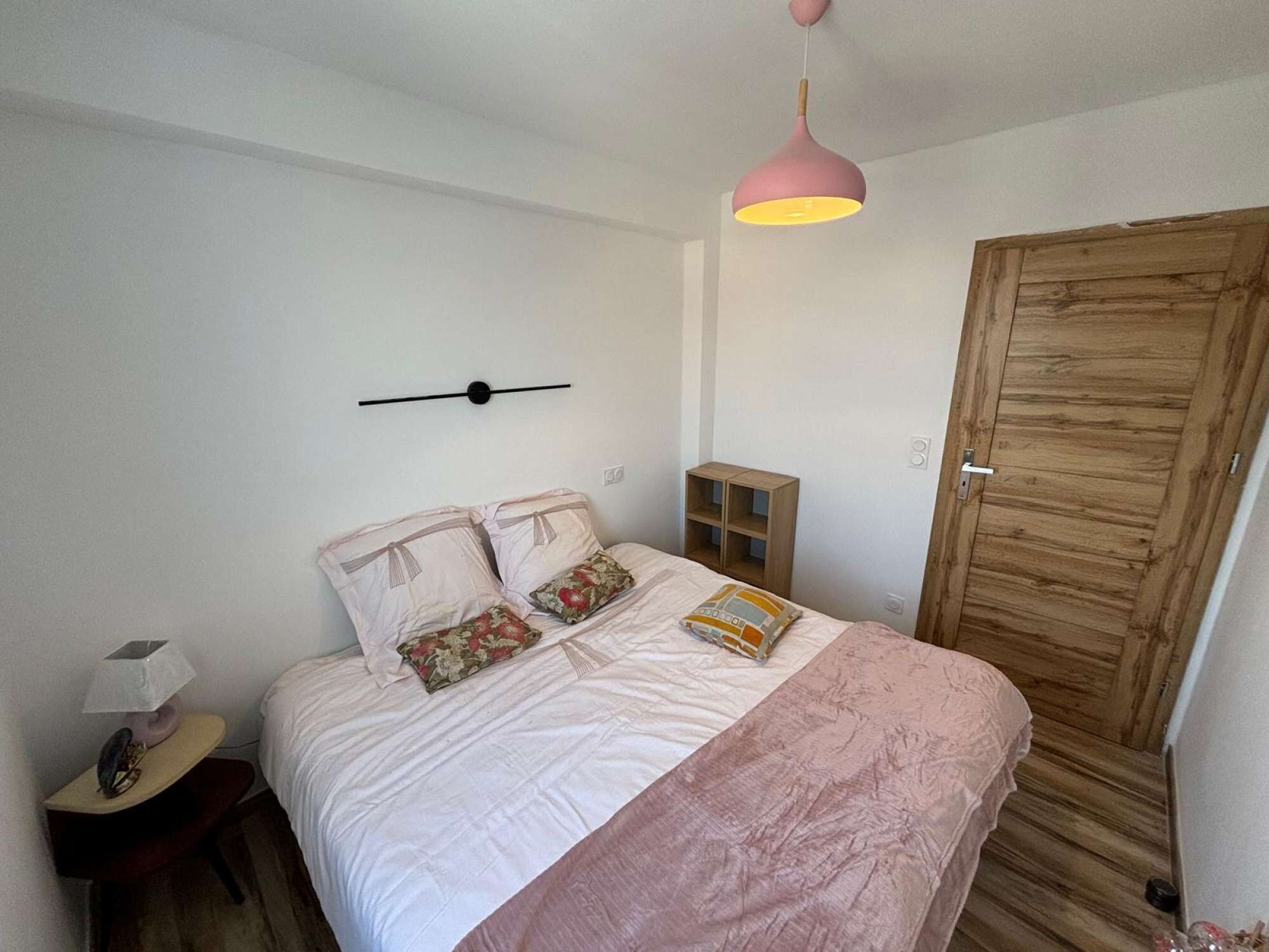 A bedroom with a bed, a bedside table, a lamp, a wooden wardrobe, and a hanging light fixture.