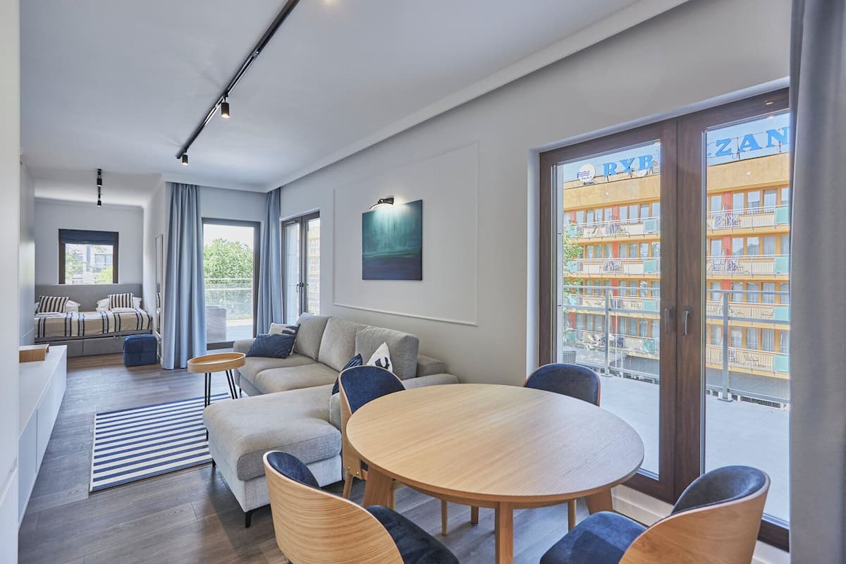 Ein modernes Hotelzimmer mit einer Sofagruppe, einem Esstisch und Blick auf ein Gebäude mit Balkonen.