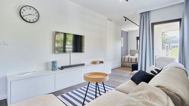 A modern living room with a flat-screen TV, a clock, a sofa, and a dining area with a striped tablecloth.