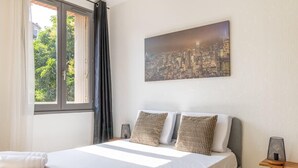 Una habitación de hotel con una cama, una pintura de paisaje urbano, una ventana con cortinas y una mesita de noche con una lámpara.