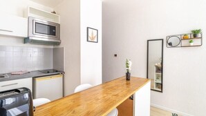 Cocina compacta con microondas, estufa y gabinetes blancos. Hay una mesa de madera con sillas, una pequeña planta en maceta y un espejo con repisas.